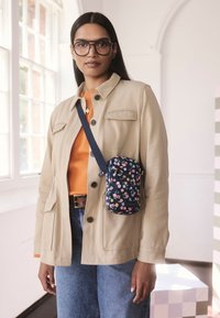 Kvinna med långt mörkt hår som bär stora glasögon, beige jacka, orange topp, blå jeans och en liten tvärgående blommig väska inomhus vid ett fönster.