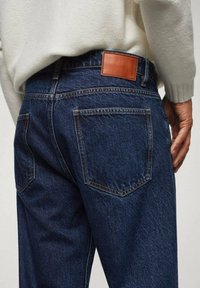 Jeans in denim blu scuro presentano una toppa in pelle sulla vita, due tasche posteriori e dettagli di cucitura visibili. Indossati con un maglione chiaro.