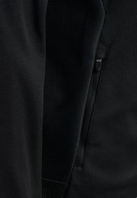 Schwarze Sportjacke mit glatter Außenseite und Netz-Innenfutter. Verfügt über eine Reißverschlusstasche und strukturierten Stoff an der Seite.