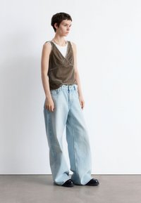 Braun, ärmelloses Oberteil aus strukturiertem Stoff über einem weißen T-Shirt; kombiniert mit hellblauen, weit geschnittenen Jeans und schwarzen Schuhen.