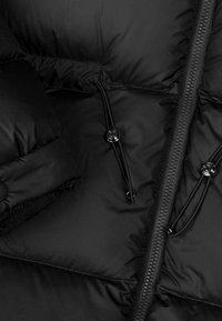 Nærbillede af sort pufferjakke med quiltet tekstur, sort lynlås, snoretræk og blanke knapper med mat finish.