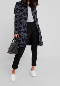 Grijze camouflagejas met zwarte accenten, knoopsluiting, gecombineerd met zwarte broek en witte instapschoenen. Zwarte handtas met gouden hardware.