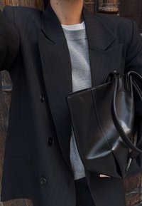 Osoba ubrana w czarną marynarkę w prążki na szarym swetrze, trzymająca dużą czarną skórzaną torbę typu tote na tle drewnianej ściany.