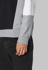 Person wearing gray, white, and black color-block sweatshirt with cuffed sleeves and black pants, standing against a plain gray background.