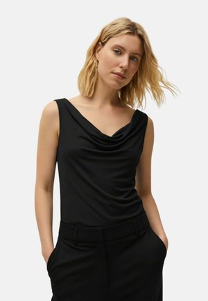 Mujer rubia con cabello hasta los hombros que lleva una blusa negra sin mangas drapeada y pantalones negros, de pie con las manos en los bolsillos.