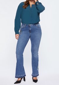 Teal blouse with button placket paired with blue flared jeans featuring beaded floral embroidery on the front, completed with black heeled shoes.