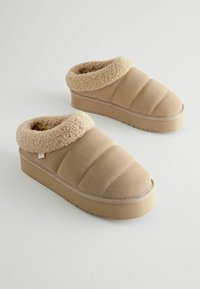 Pantoufles beiges à enfiler avec un col en crème pelucheux, un design rembourré et une semelle en caoutchouc texturé, présentant une étiquette de logo discrète sur le côté.