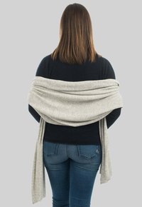 Lys grå kappe draperet over en sort sweater, der har en blød tekstur og et flydende design, båret af en person, der vender væk, med blå jeans synlige.