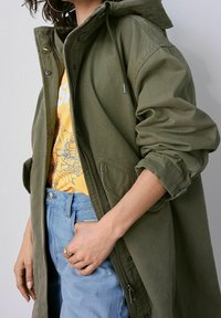 Personne portant une veste vert olive par-dessus un t-shirt graphique jaune et un jean bleu clair, une main dans la poche du jean.