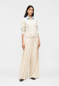 Γυναίκα που στέκεται φορώντας ένα ανοιχτό γαλάζιο πουκάμισο με γιακά, κρεμ ζακέτα με κουμπιά, φαρδιά μπεζ πλισέ παντελόνια και μαύρες μπαλαρίνες με χρυσά καρφιά.