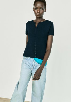 Femme portant un cardigan bleu marine à manches courtes boutonné sur un haut turquoise et un jean large bleu clair, debout devant un fond uni.