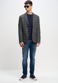 Šedý kostkovaný blejzr se dvěma knoflíky přes tmavě modrý pletený svetr, kombinovaný s modrými džíny a tmavomodrými teniskami. Model má na sobě tmavé sluneční brýle.