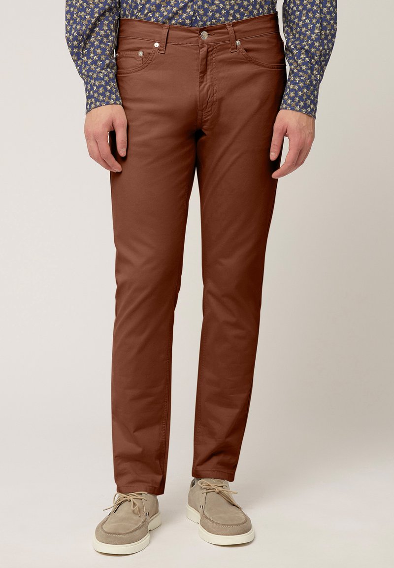 Braune Baumwollhose mit schmaler Passform, die eine glatte Textur, ein Fünf-Taschen-Design und metallische Beschläge aufweist. Getragen mit hellen Schuhen.