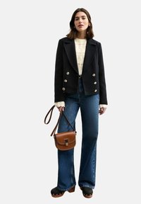 Veste noire croisée avec des boutons argentés, pull en tricot crème, jeans évasés taille haute, sac à main marron et sabots noirs.