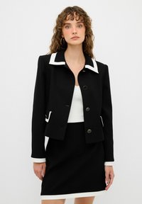 Zwarte cropped jas met witte kraag en accenten, voorzien van gouden knopen. Gecombineerd met een zwarte rok en een strakke witte top. Soepele textuur.