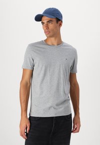 Tricou gri cu mânecă scurtă, cu guler rotund și un logo mic pe pieptul stâng. Purtați cu o șapcă de baseball albastră, asortată cu blugi negri.