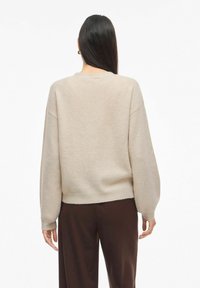 Žena s dlouhými tmavými vlasy má na sobě béžový svetr s balónovými rukávy a tmavě hnědé kalhoty, zobrazená ze zadu na bílém pozadí.