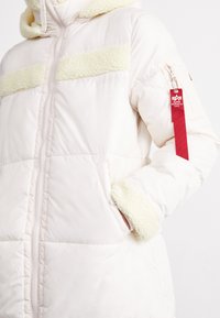 Biała kurtka puchowa z miękkim żółtym paskiem z sztucznego futra, zamek błyskawiczny, kieszenie boczne i czerwona metka na rękawie. Teksturowany detal przy mankietach.