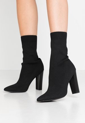 Bottes chaussettes noires à talons hauts portées sur des jambes nues, avec des poignets côtelés et des bouts pointus sur une surface lisse et réfléchissante.