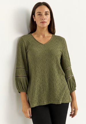 Woman with straight brown hair wearing olive green textured V-neck blouse with lace insert sleeves and black pants, standing against white wall.