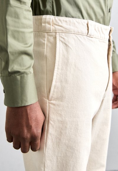 Pantaloni din bumbac bej cu un finisaj texturat, cu buzunare laterale și fermoar, asortați cu o cămașă verde cu nasturi.