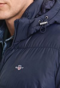 Jūras spēku pūkainā jaka ar augstu apkakli, ar rāvējslēdzēju priekšpusē un logo detaļu uz krūtīm; gluda, spīdīga tekstūra. Ietver regulējamu auklu.