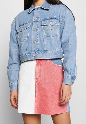 Vrouw draagt een cropped lichtblauw spijkerjasje en een high-waisted rok met witte, rode en blauwe kleurvlakken, staand met handen langs het lichaam.