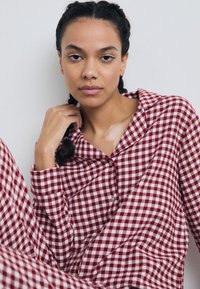 Kvinde med fletninger iført rød- og hvidternet pyjamas, siddende mod en hvid væg, med hånden nær halsen.