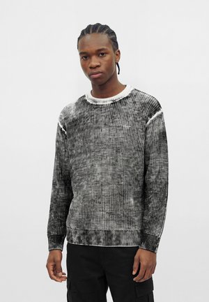 Jeune homme aux cheveux tressés portant un pull en maille texturée noir et blanc et un pantalon noir sur un fond blanc uni.