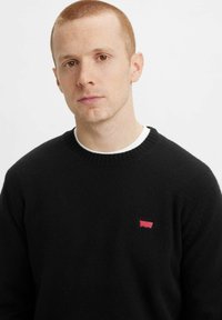 Maglione nero realizzato in materiale morbido, con un collo a girocollo a coste e un piccolo logo rosso sul lato sinistro del petto. Design semplice e classico.
