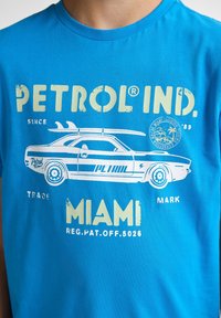 Camiseta de algodón azul con un gráfico de un coche vintage y el texto "PETROL IND." en amarillo, con un diseño de tabla de surf y referencia a Miami.