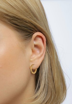 Boucle d'oreille en or avec une surface texturée, présentant une forme circulaire et une finition polie, portée sur des cheveux légers et de longueur moyenne.