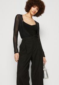 Black sheer long-sleeve top over a black tank, paired with high-waisted, textured black trousers. The model holds a silver handbag.