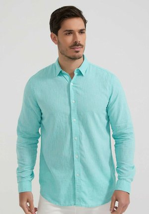 Homme aux cheveux courts et foncés portant une chemise bleue claire à manches longues boutonnée, debout devant un fond clair uni.