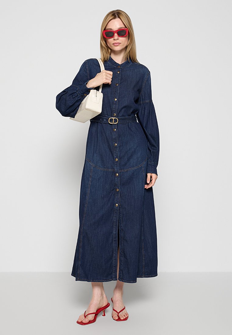 Dżinsowa sukienka z długim rękawem, zapinana na guziki z przodu, z talią ściągniętą paskiem. Modelka nosi czerwone okulary przeciwsłoneczne i trzyma małą białą torebkę.