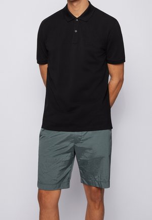 Mann trägt schwarzes Poloshirt mit kurzen Ärmeln und glänzende graue knielange Shorts, steht mit den Händen hinter dem Rücken vor einem einfarbigen Hintergrund.