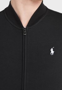 Svart zip-up jacka med ribbad krage och en liten vit polo-spelare-logotyp på vänster bröst.