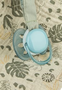 Light blue pacifier with a round shape, silicone material, a flexible ring, and a cloth strap, set against a patterned fabric background.