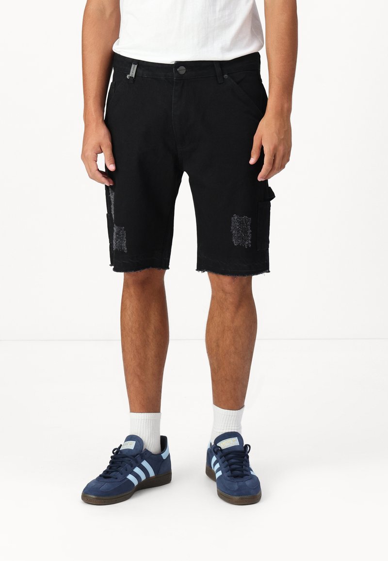 Pantalones cortos de mezclilla negra con dobladillos deshilachados, que presentan parches desgastados y bolsillos laterales, combinados con zapatillas deportivas azules y calcetines blancos.