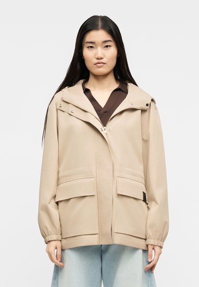 Femme aux longs cheveux noirs portant une veste beige zippée avec poches à rabat sur un haut marron et un jean large bleu clair, debout devant un fond blanc.