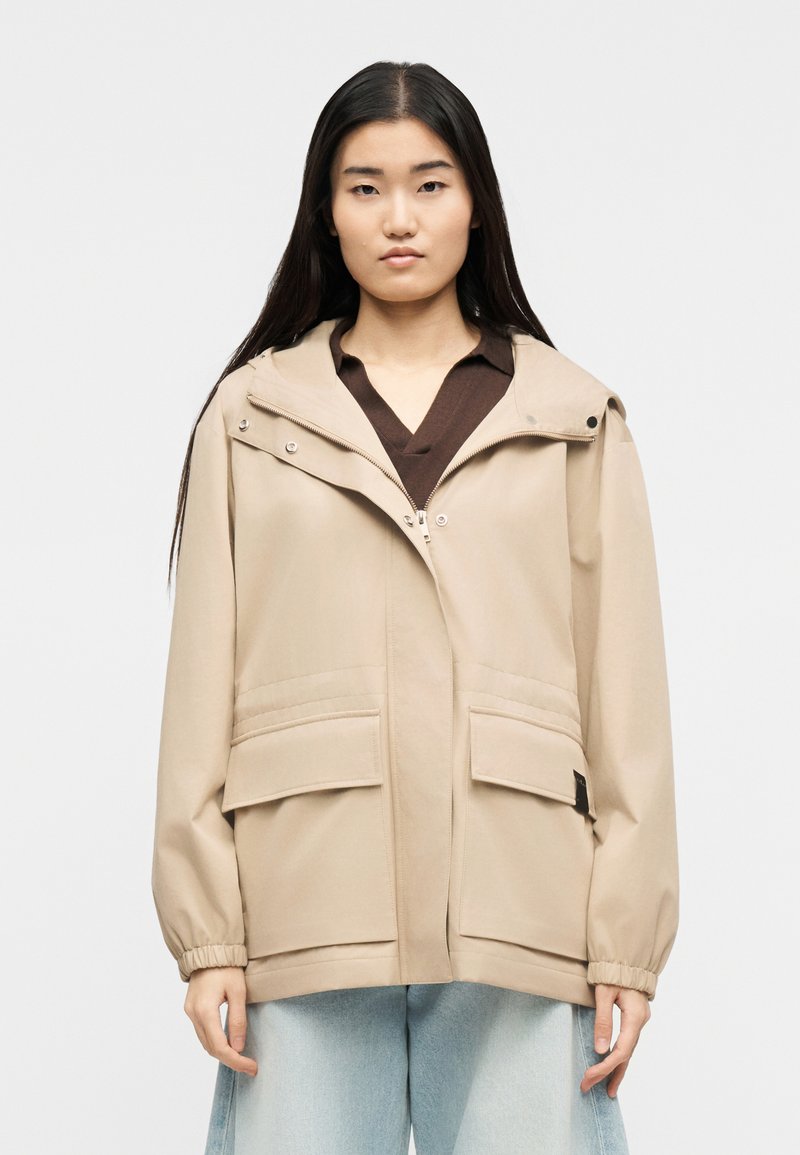 Γυναίκα με μακριά σκούρα μαλλιά φοράει μπεζ ζακέτα με φερμουάρ και τσέπες με καπάκι πάνω από καφέ μπλουζάκι και ανοιχτό μπλε τζιν με φαρδιά μπατζάκια, στέκεται μπροστά σε λευκό φόντο.