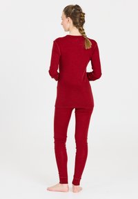 Vrouw met gevlochten haar, gekleed in een rood langermig shirt en bijpassende legging, staat op blote voeten met handen in de zij, met de rug naar de camera.