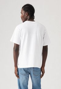 Mand med flettet hår iført en almindelig hvid T-shirt og blå jeans, stående med ryggen til mod en lys grå baggrund.