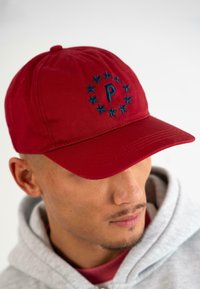 Căciulă de baseball roșie din material textil, cu litera albastră "P" brodată, înconjurată de stele. Textură moale, cu boruș curbat și spate ajustabil.