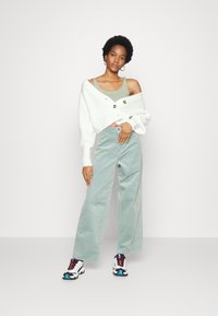 Calças largas de corduroy verde-claro combinadas com um cardigan oversized branco de malha com botões grandes, e uma t-shirt de alças menta por baixo. Ténis com detalhes em preto, azul e vermelho.