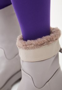 Botas impermeables de tobillo en gris claro con un puño ancho que presenta un forro de piel suave beige. Leggings morados visibles encima de la bota.