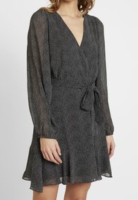 Svart omlotklänning med vita prickar, lång ärm, V-ringning och en knytning i midjan. Lätt och flödande tyg, samlad vid ärmsluten.