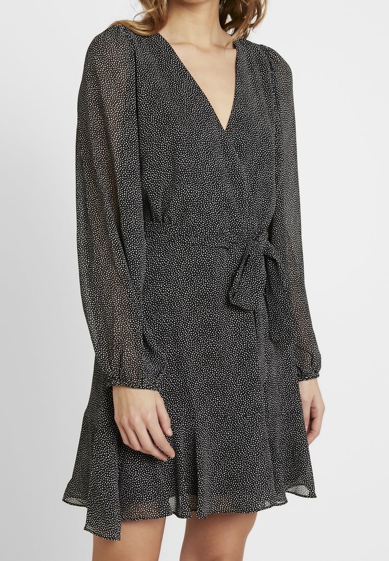 Svart omlotklänning med vita prickar, lång ärm, V-ringning och en knytning i midjan. Lätt och flödande tyg, samlad vid ärmsluten.