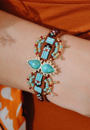 Türkis- und goldfarbenes Perlenarmband mit einem Lederband. Es weist intricate Muster, goldene Akzente und tropfenförmige Steine auf.