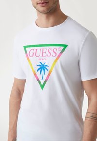 Hvid bomulds T-shirt med et trekantet grafisk design i gr�øn, pink og gul, med teksten "GUESS BEACH" og en illustration af et palmetræ.